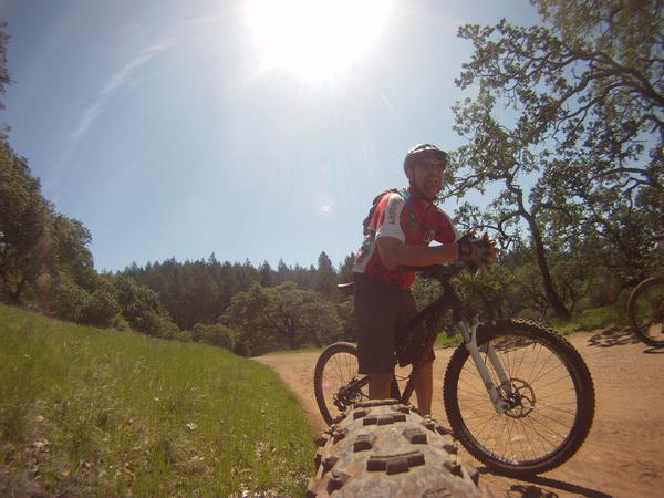 Santa Cruz Tallboy: A mountain biker wearing a helmet and red jersey stands next to his bike on a sunny day, surrounded by green trees and a dirt trail. The sun is bright in the sky, casting a warm glow over the landscape.