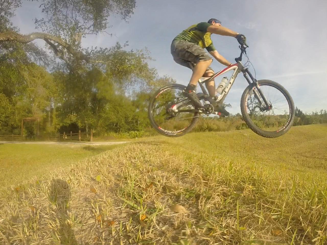 A cyclist performing a jump on a mountain bike over a grassy area, with trees and a pathway in the background on a sunny day. Nocatee mountain bike trail.