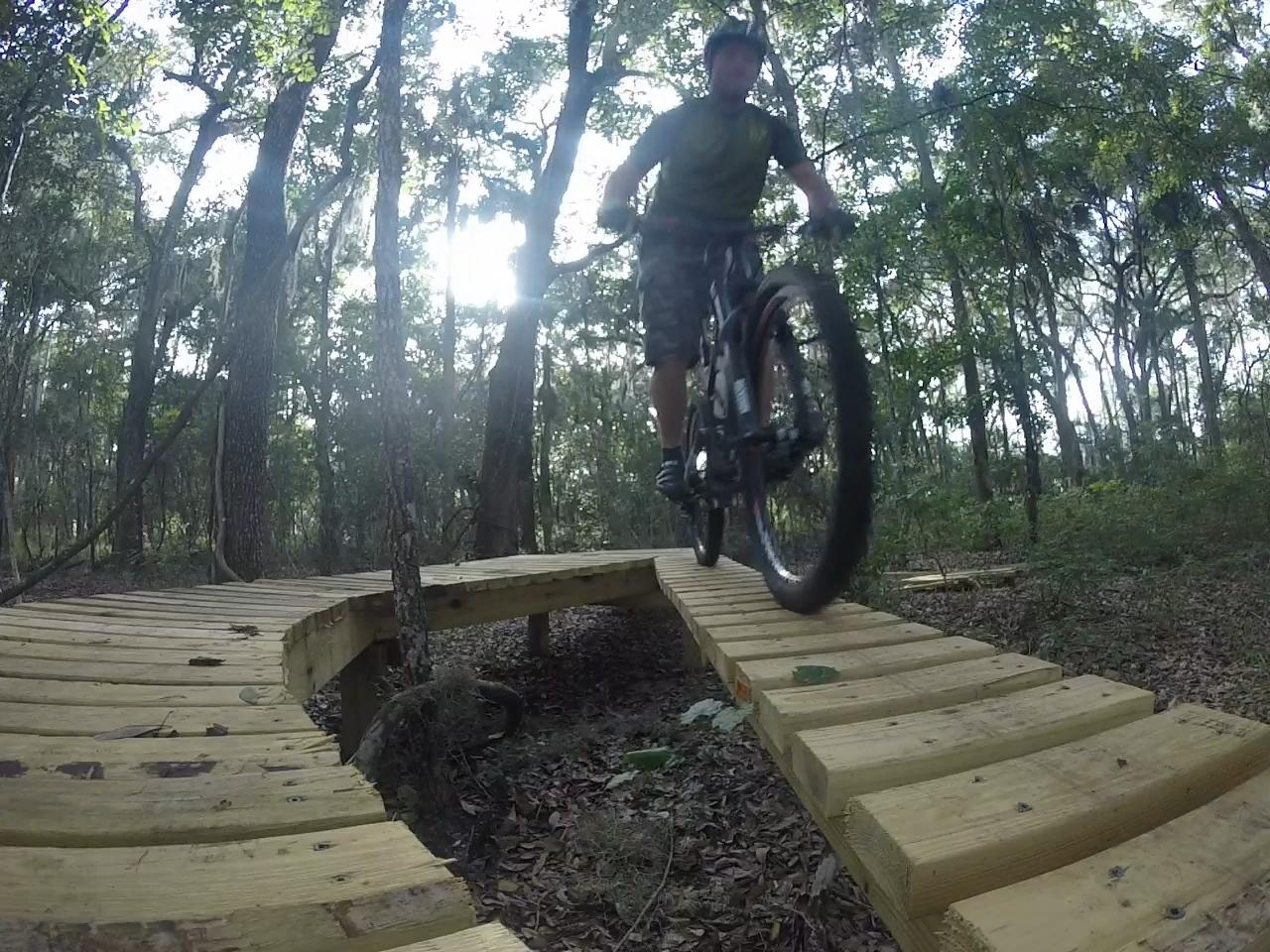 A cyclist riding a mountain bike over a wooden bridge on a forest trail, surrounded by tall trees and dappled sunlight. Nocatee mountain bike trail.