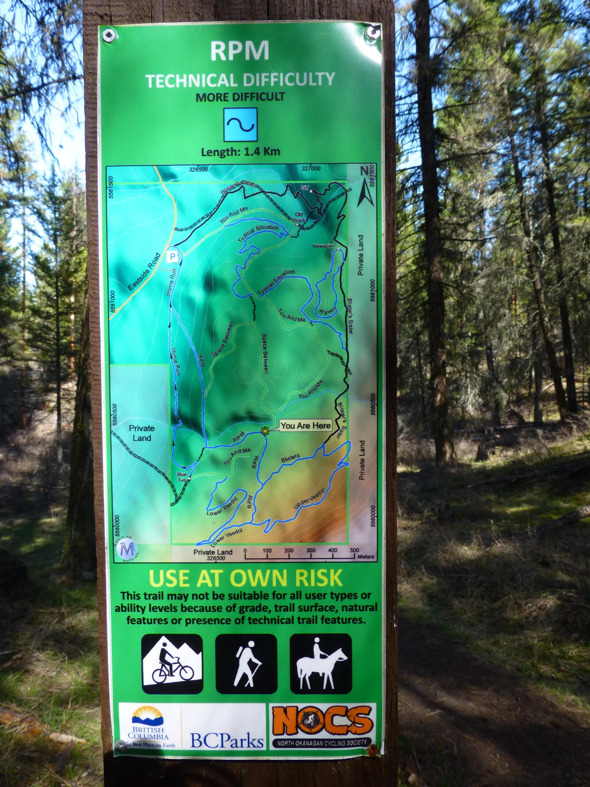 Sign showing the RPM trail map with technical difficulty indicated as "more difficult." It features a detailed map outlining a 1.4 km trail, noting "You Are Here" and surrounding landmarks. The sign includes a caution statement advising users to "use at own risk" due to variable trail conditions. Icons represent different trail users: cyclists, hikers, and horseback riders. RPM mountain bike trail.