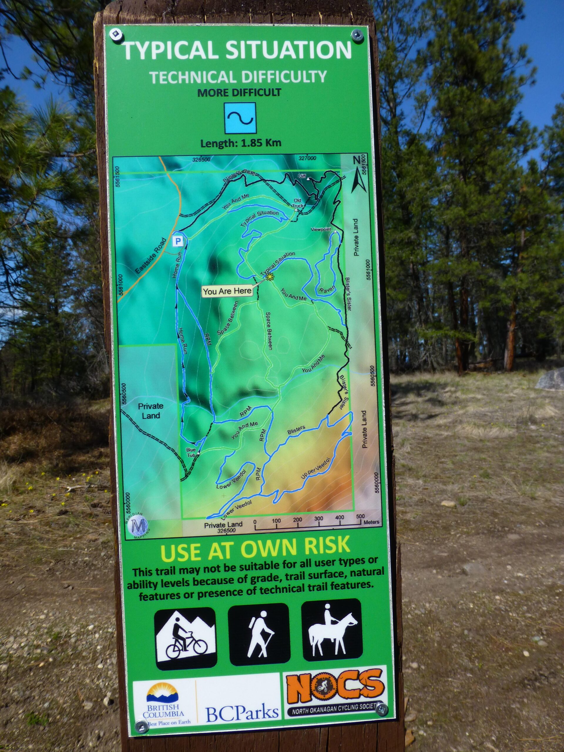 A trail map sign indicating the technical difficulty of a trail labeled "Typical Situation." The map shows a length of 1.85 km with a marked location reading "You Are Here." It features warnings to use the trail at one's own risk and notes that the trail may not be suitable for all user types. Icons representing biking, hiking, and horseback riding are included at the bottom alongside logos for BC Parks and the North Okanagan Cycling Society. The sign is set against a backdrop of trees. Typical Situation mountain bike trail.