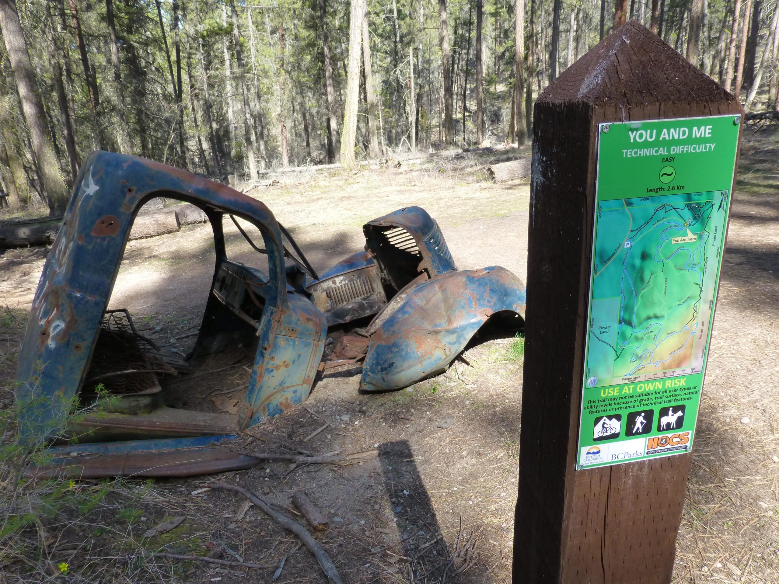 An old, rusted car shell lies on the ground surrounded by trees in a forested area, next to a wooden sign. The sign displays a trail map labeled "You and Me," indicating an easy technical difficulty with a length of 2.6 km, and includes warnings to use the trail at one