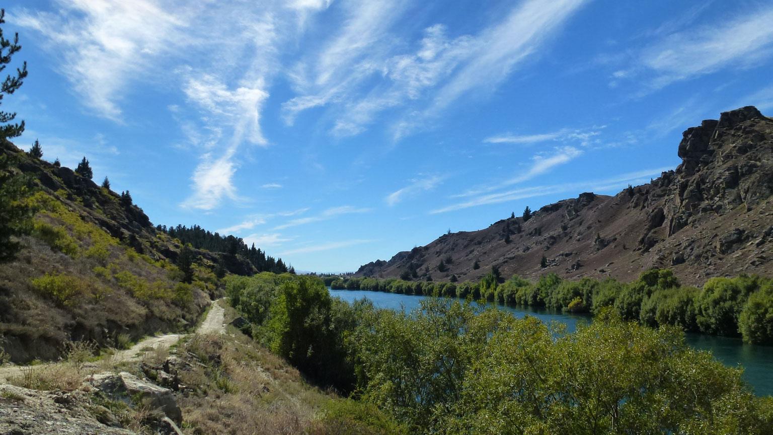 A serene landscape featuring a winding river bordered by lush greenery and rugged hills under a clear blue sky with scattered wispy clouds. A dirt path runs alongside the river, inviting exploration of the natural surroundings. Roxburgh Gorge Trail mountain bike trail.