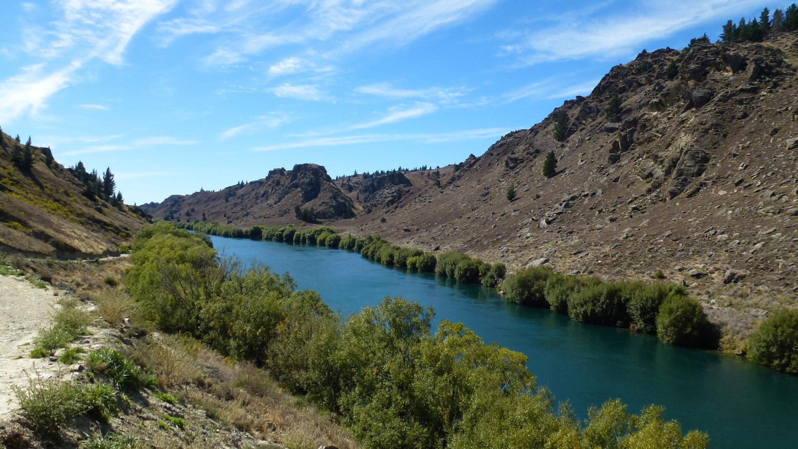 A serene river flows through a rugged landscape, flanked by rocky hills and green vegetation. The clear blue sky is dotted with wispy clouds, while shrubs and trees line the riverbanks, enhancing the natural beauty of the scene. The image captures a peaceful outdoor environment, perfect for nature lovers. Roxburgh Gorge Trail mountain bike trail.