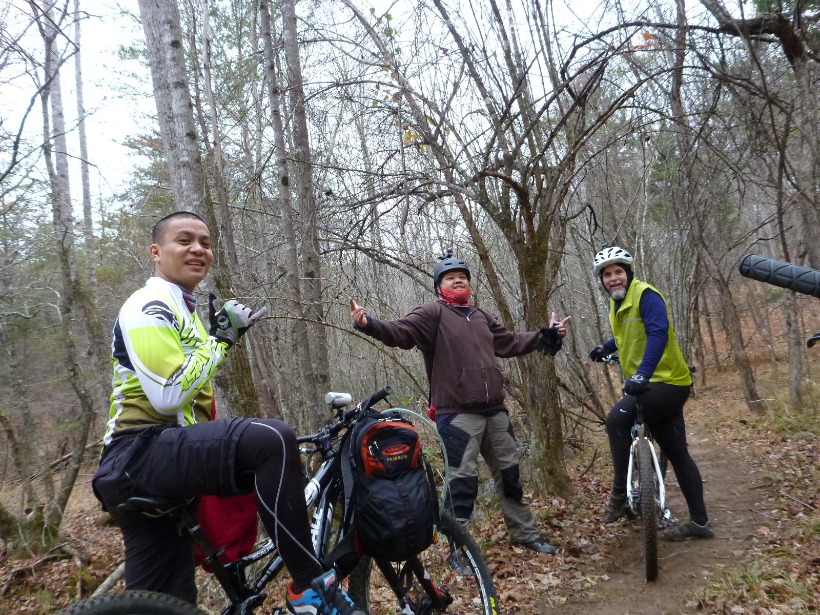 A group of three mountain bikers smiling and posing for a photo on a wooded trail. The first rider is wearing a bright green and white cycling jersey while sitting on a bike. The second rider, dressed in a brown jacket and red scarf, is gesturing with both hands. The third rider, in a green vest and black gloves, is standing next to his bike. The background features tall, bare trees and a leaf-strewn path, indicating a cool, autumn day. Mountain Laurel Trails mountain bike trail.
