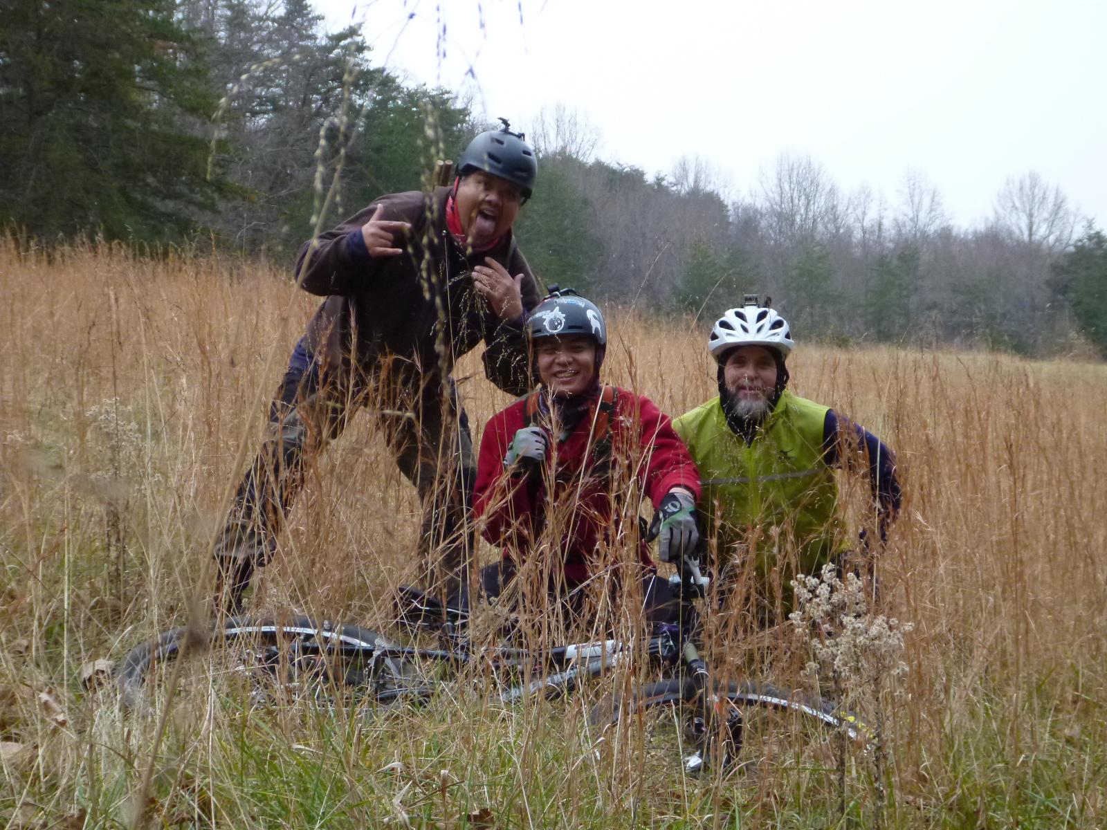 Three people wearing helmets and biking gear are posing playfully in a field of tall grasses. One person is making a silly face, while another is crouching in front of a mountain bike. The background features trees and a cloudy sky, suggesting an outdoor setting. Mountain Laurel Trails mountain bike trail.