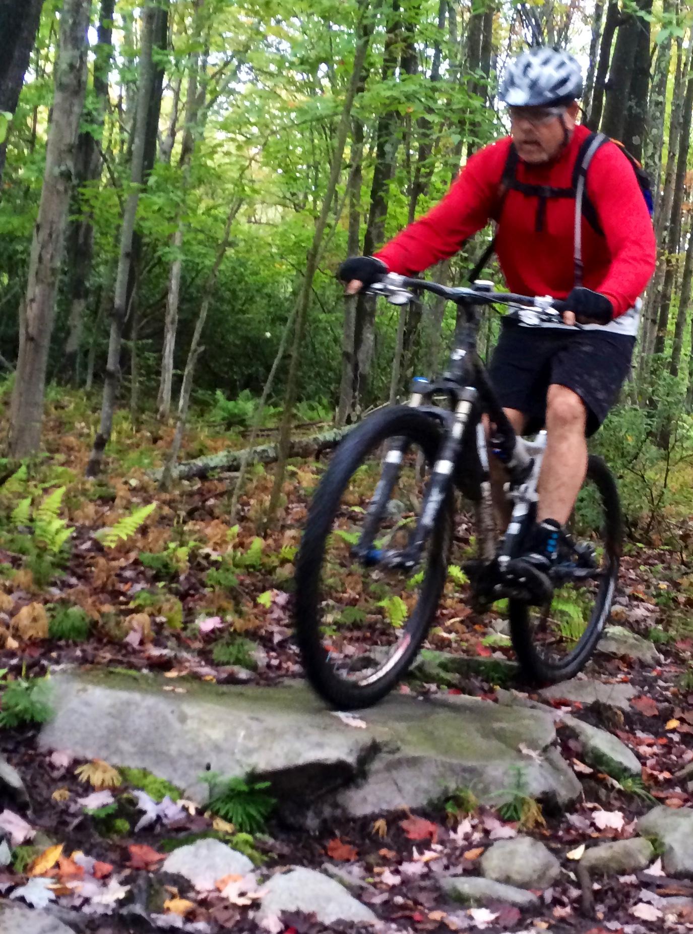A mountain biker in a red long-sleeve shirt rides over a rocky trail in a lush, green forest during autumn, with fallen leaves scattered on the ground. CVI Trails mountain bike trail.