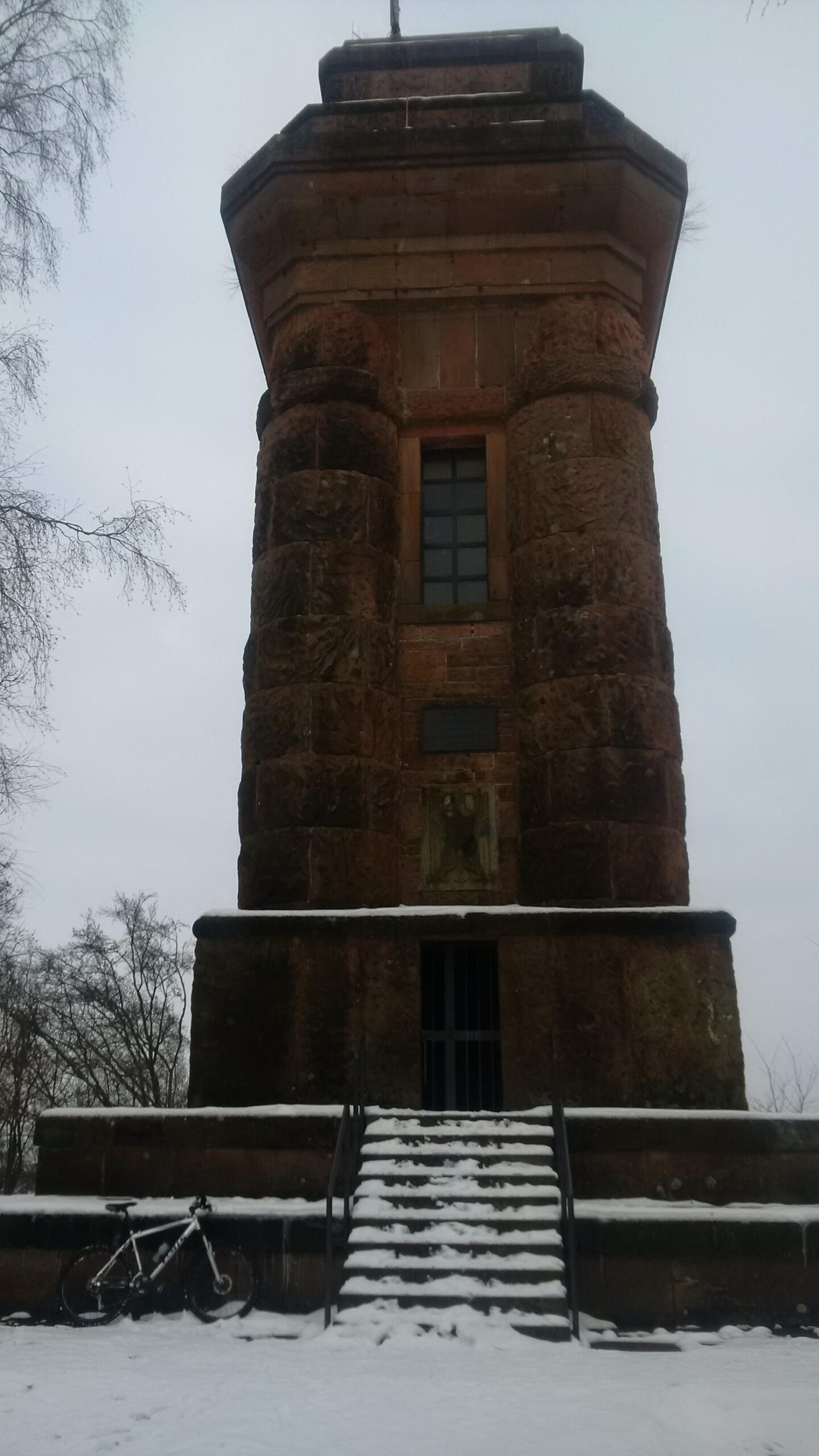 A tall, historic stone tower stands prominently in a snowy landscape. The structure features a rectangular base, with a small staircase leading up to a gated entrance. Snow covers the ground around the tower, while a bicycle rests against the lower wall. In the background, branches of bare trees are visible against a gray sky. Tower Trail mountain bike trail.