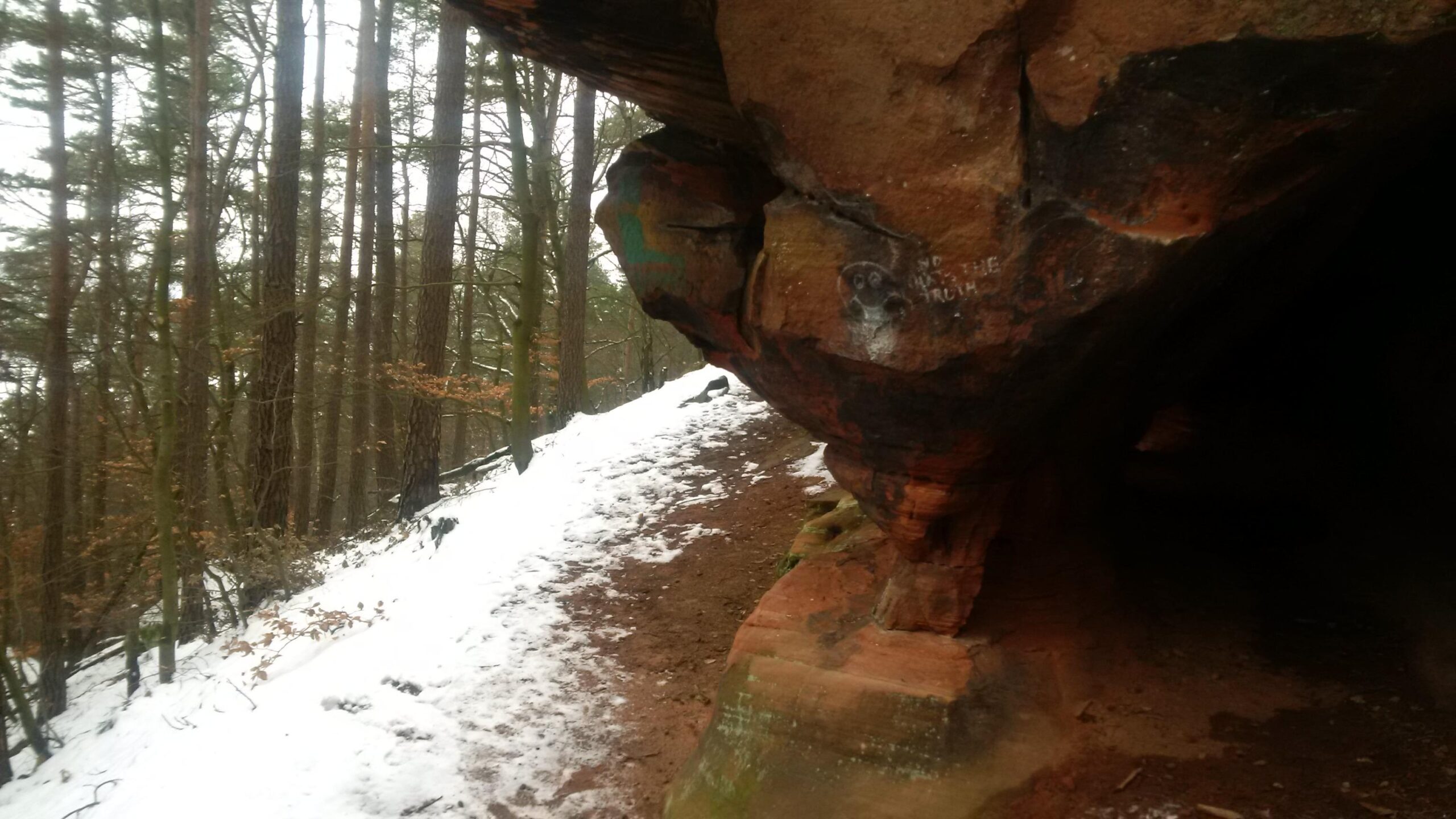 A rocky overhang beside a snow-dusted path, surrounded by tall trees in a forested area. The ground features a mix of dirt and some remaining patches of snow. The rock surface has graffiti and markings, adding a touch of human presence to the natural setting. Tower Trail mountain bike trail.
