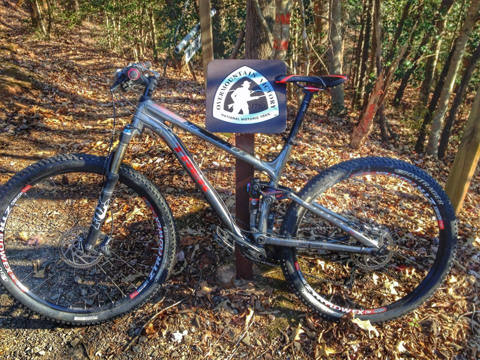 Trek Fuel EX 8 29: A mountain bike resting beside a trail sign for the Overmountain Victory National Historic Trail, surrounded by autumn leaves and trees in the background.