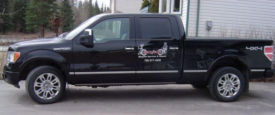 A black Ford F-150 truck parked beside a building, featuring a logo and text for a bicycle service and repair business. The landscape includes trees in the background.