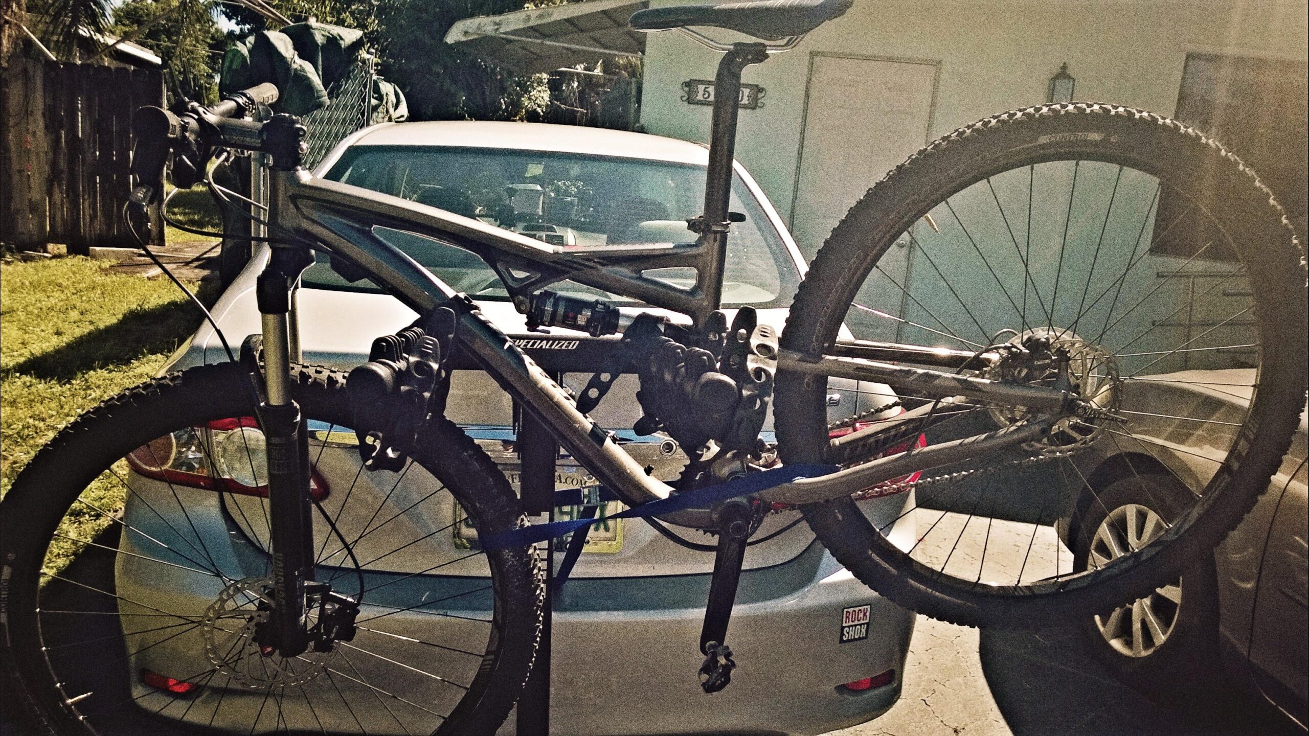 Specialized Camber: A mountain bike is mounted on a bike rack, positioned behind a silver sedan parked in a residential area. The bike features a gray frame with suspension components visible, and it displays knobby tires suitable for off-road riding. In the background, there are elements of a garden and a house. The setting is sunny with clear, blue skies.