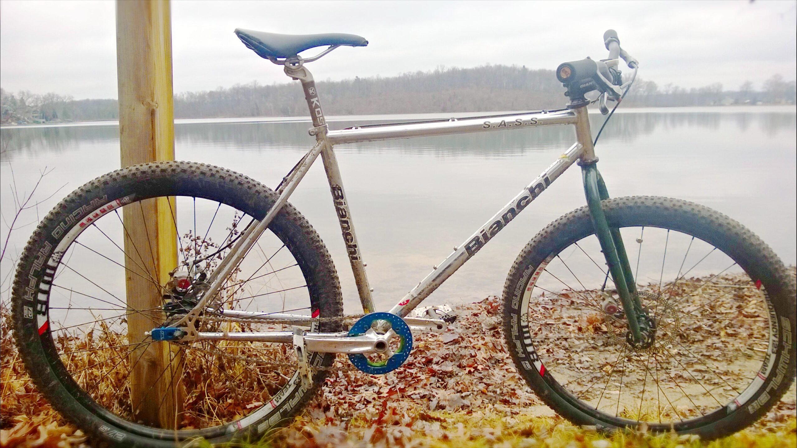 Bianchi S.A.S.S.: A silver Bianchi mountain bike with a blue chainring stands near a calm lake. The bike is partially muddy, indicating recent use, and is positioned next to a wooden post. Behind the bike, the lake is surrounded by trees under a cloudy sky, with fallen leaves scattered on the ground.