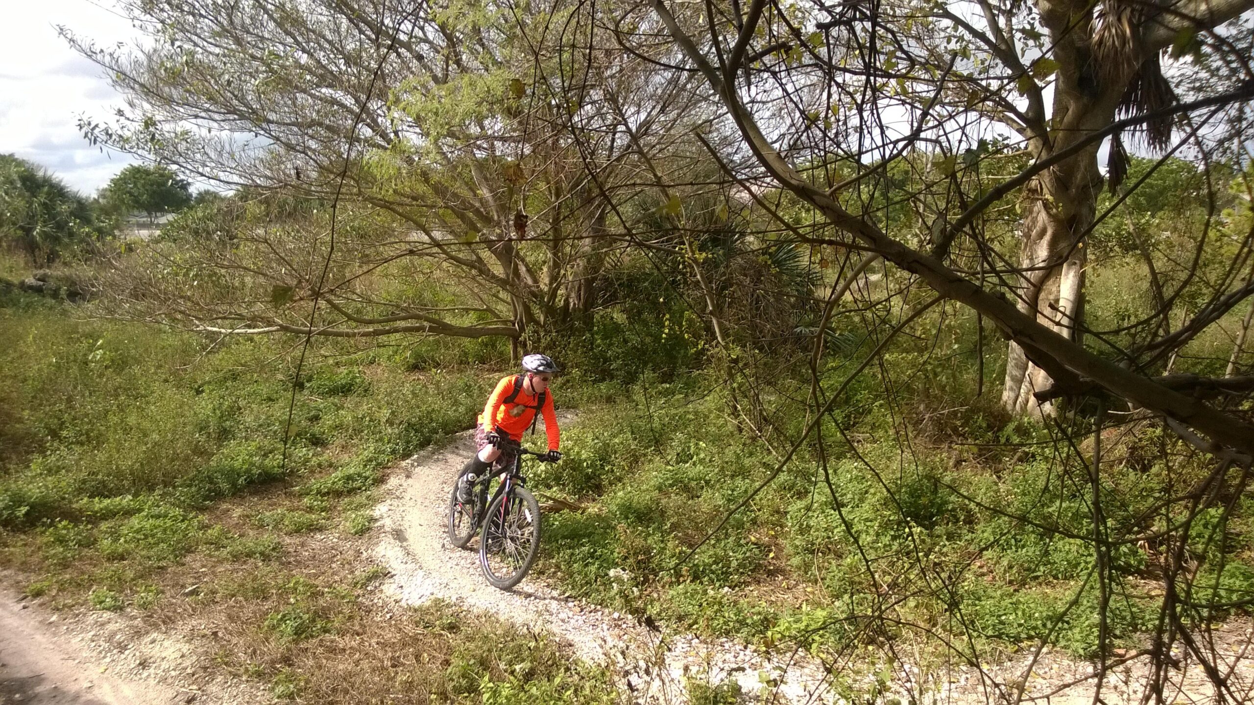 A cyclist in a bright orange jacket rides a mountain bike along a winding gravel path surrounded by lush greenery and sparse trees. The scene is set in a natural outdoor environment with patches of sunlight filtering through the branches. Quiet Waters Park mountain bike trail.