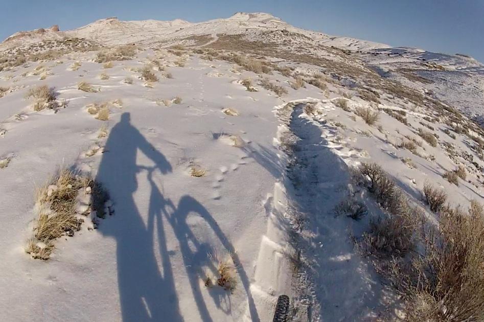 A shadow of a cyclist riding on a snow-covered trail, surrounded by sparse vegetation and hills in the background. The scene captures the serene beauty of winter biking in a natural landscape. Tnt mountain bike trail.