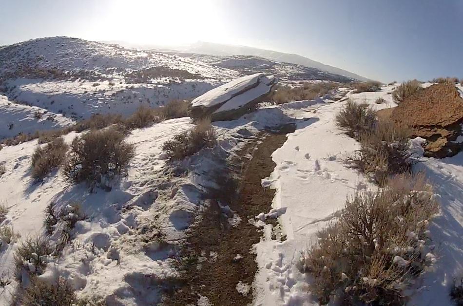 A snowy landscape featuring a narrow dirt path winding through shrubs and rocks, with rolling hills in the background. The sun is shining brightly, illuminating the scene. Tnt mountain bike trail.