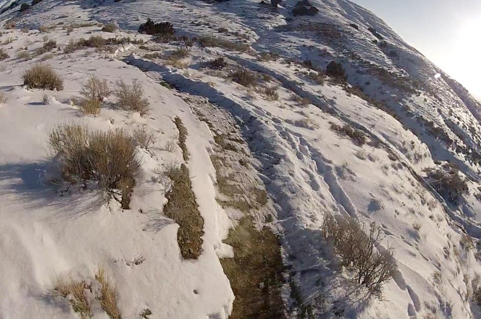 A winding dirt path leads through a snow-covered landscape, surrounded by patches of grass and sparse shrubs. The scene is illuminated by sunlight, casting soft shadows on the white snow and highlighting the contours of the hilly terrain. Tnt mountain bike trail.