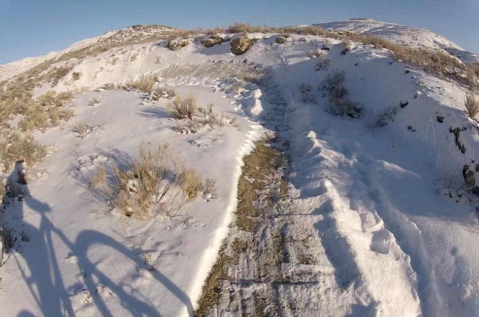 Alt text: A snowy trail winding through a hilly landscape, with patches of bare ground visible. Sunlight casts shadows on the path, and sparse vegetation is seen along the edges of the trail. Tnt mountain bike trail.