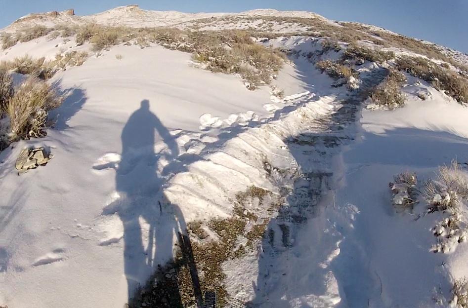 A cyclist's shadow is visible on a snowy trail winding through the hills, with patches of grass peeking through the snow. The scene captures a bright, sunny day in a winter landscape, highlighting the mix of snow-covered ground and rugged terrain. Tnt mountain bike trail.