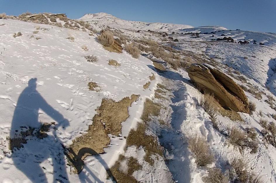A snowy mountain trail with a bicycle and the shadow of the rider visible, surrounded by snow-covered hills and rocky formations under a clear blue sky. Tnt mountain bike trail.