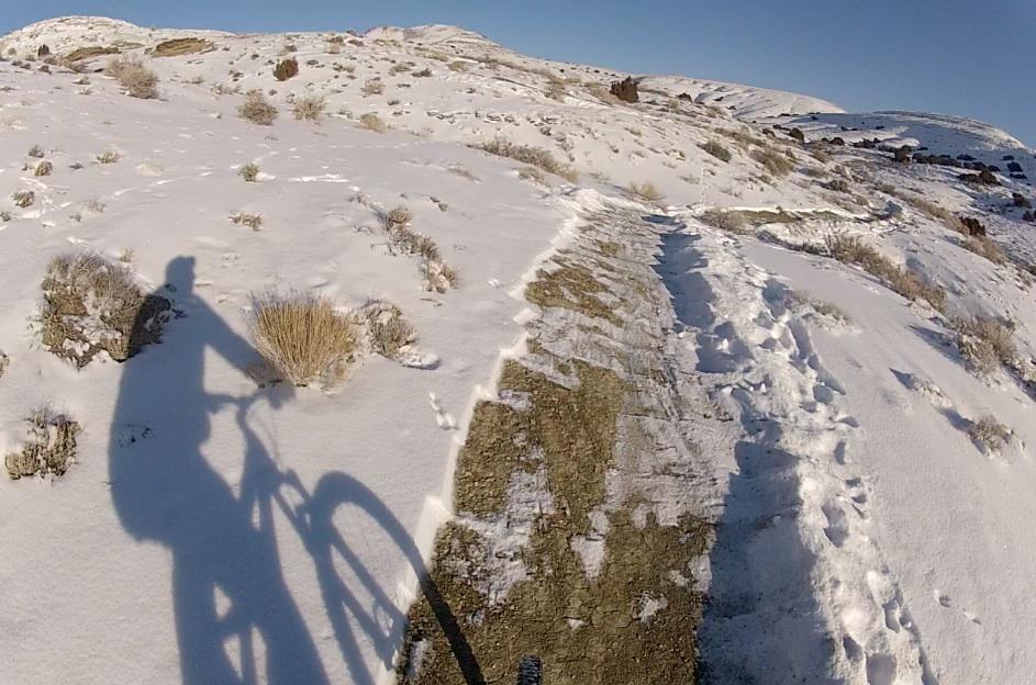 A shadow of a mountain biker on a snow-covered trail, with rugged hills and sparse vegetation in the background. The pathway is partially visible, showing a mix of snowy and dirt surfaces, suggesting a winter outdoor adventure. Tnt mountain bike trail.