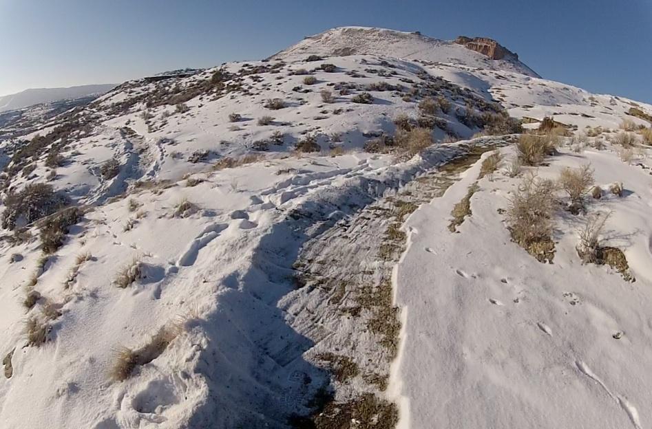 A snow-covered hillside with a clear blue sky, featuring a narrow trail winding through the snow and sparse vegetation. In the background, a rocky outcrop rises above the landscape, contrasting with the white snow. Soft shadows are cast across the terrain. Tnt mountain bike trail.