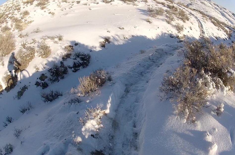 A snowy trail winding through rugged terrain, with patches of shrubs visible along the path. A shadow of a cyclist is cast on the snow as they ride along the trail, highlighting the winter landscape. The scene captures a peaceful yet adventurous outdoor atmosphere. Tnt mountain bike trail.