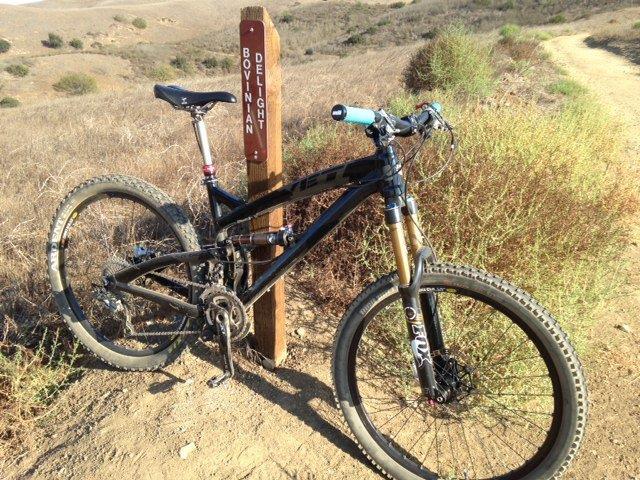 Yeti SB66: A black mountain bike parked next to a wooden trail sign labeled "Bovinan Delight," surrounded by arid terrain and sparse vegetation. The path leads through a hilly landscape.