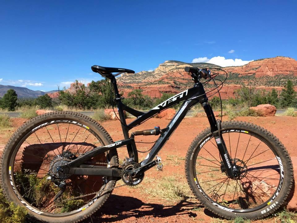 Yeti SB-75: A black mountain bike placed on a red dirt trail, with rocky terrain and green vegetation in the background. The scene features blue skies and distant hills, showcasing a vibrant outdoor setting.