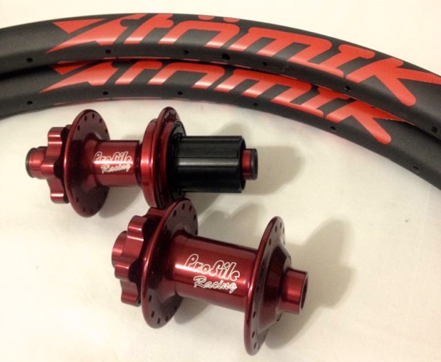 Two red bicycle hubs from Profile Racing, featuring detailed branding, positioned in front of two black bicycle rims with "Atomic" written prominently in red.