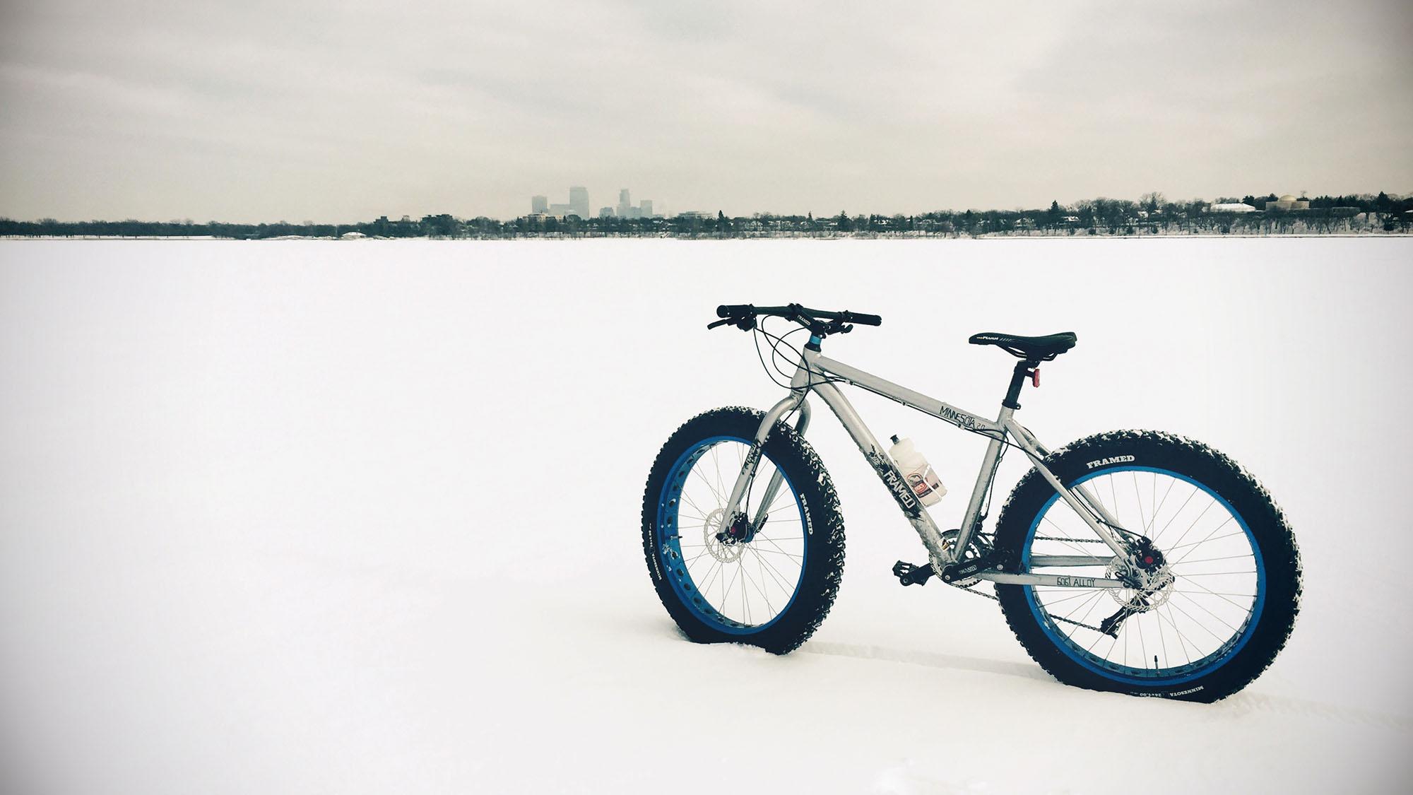 Framed Minnesota 2.0: A fat tire bicycle stands on a snowy landscape, with a city skyline visible in the background. The bike is silver with blue accents and has a water bottle mounted on the frame. The scene is overcast, emphasizing the vast white expanse of snow.