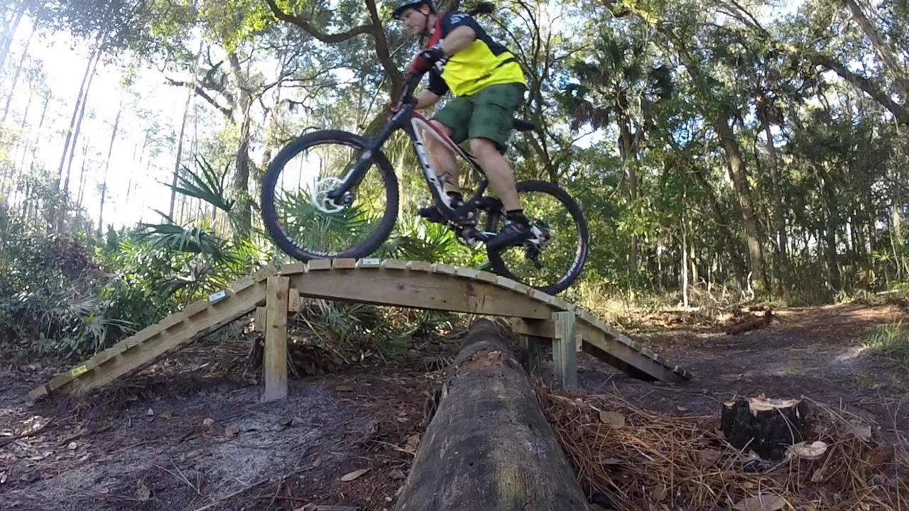 A mountain biker in a bright yellow and black jersey rides over a wooden ramp bridge in a forested area, with tall trees and lush green vegetation in the background. Nocatee mountain bike trail.