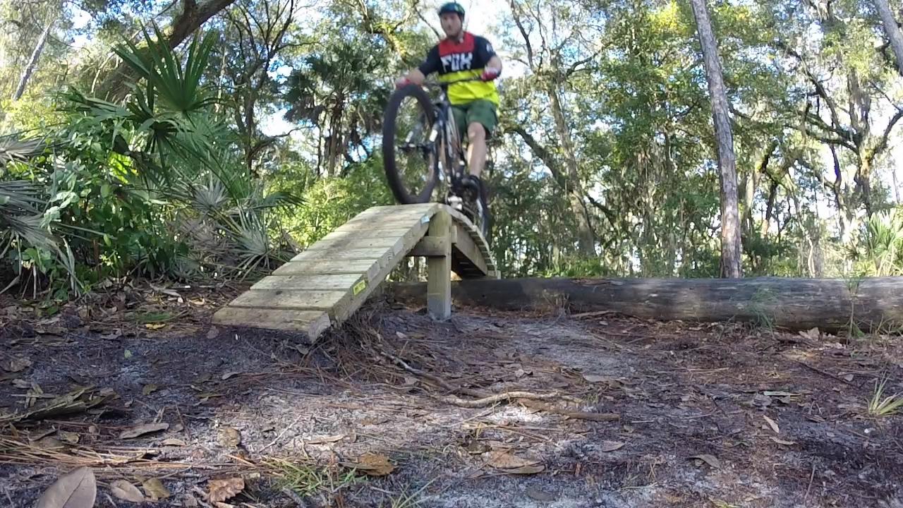 A mountain biker performing a jump over a wooden ramp on a forest trail, surrounded by greenery and tall trees. The biker is wearing a helmet and colorful cycling gear, with the front wheel lifted off the ramp. The ground is covered in leaves and dirt, indicating an outdoor cycling environment. Nocatee mountain bike trail.