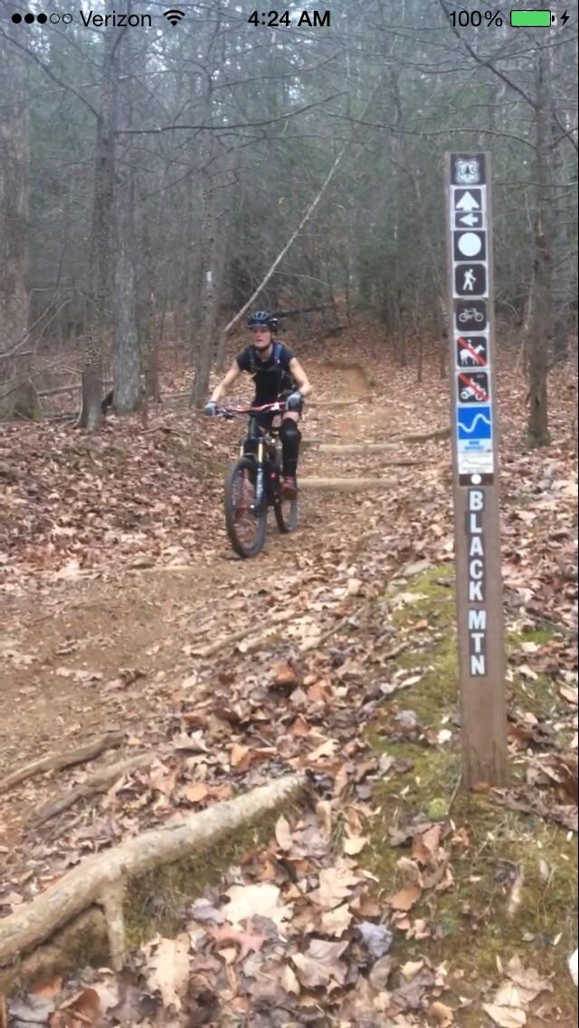 A mountain biker rides along a dirt trail surrounded by trees in a forested area. A trail sign marked "BLACK MTN" stands to the right, indicating various activities such as hiking and mountain biking. The ground is covered with fallen leaves and there are tree roots visible along the path. Black Mountain mountain bike trail.