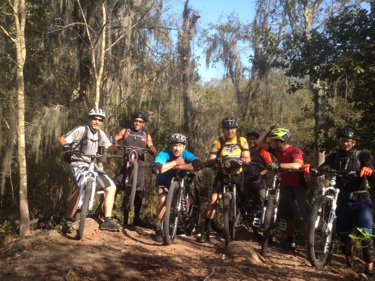 Group of six mountain bikers posing together on a wooded trail, surrounded by trees with Spanish moss. They are wearing helmets and biking gear, and some bikes are resting alongside them. The setting is bright and sunny, with green foliage in the background. Alafia River State Park mountain bike trail.