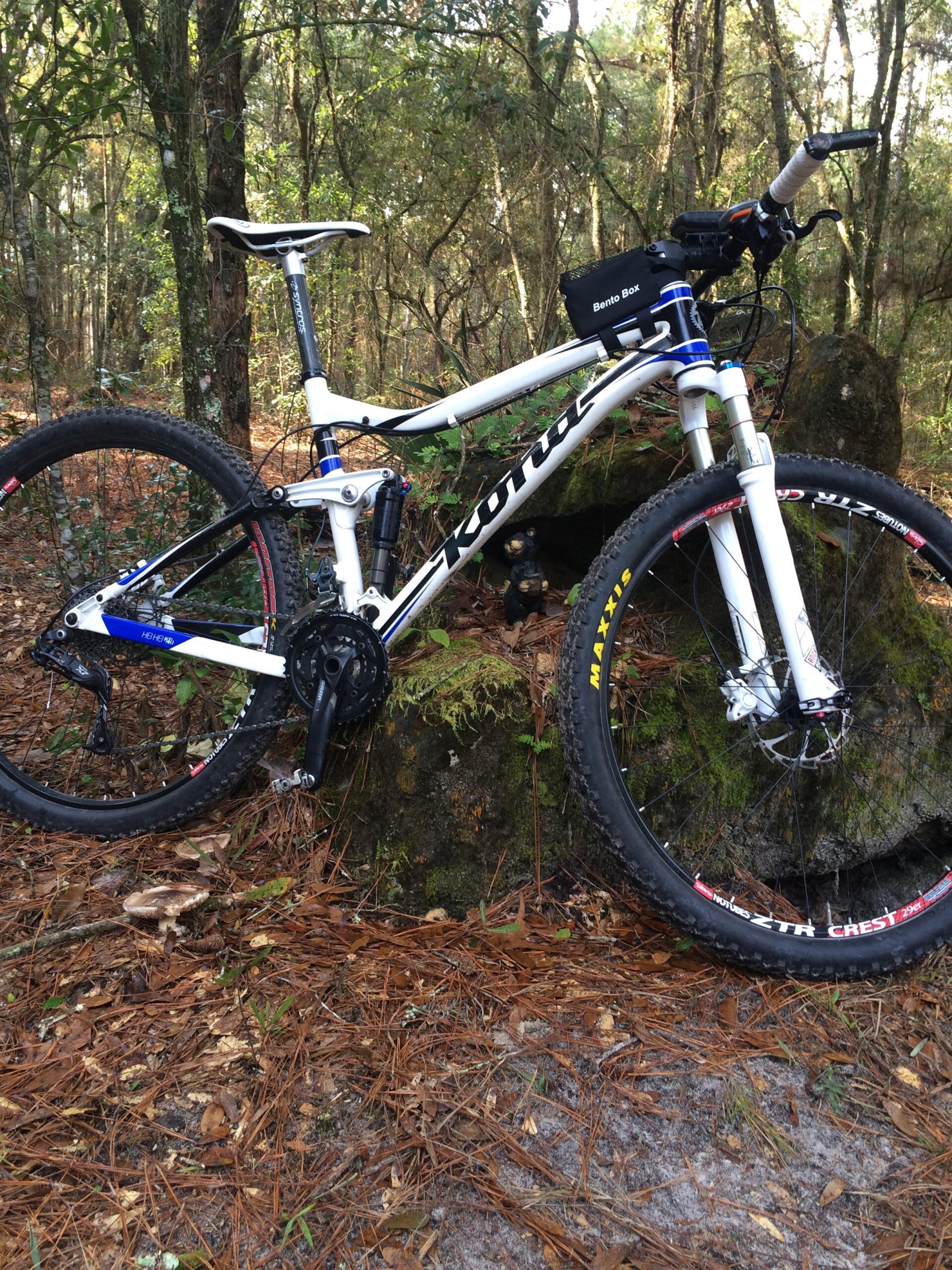 A white mountain bike with blue accents resting against a moss-covered rock in a wooded area, surrounded by pine needles and small plants. The bike features a full suspension, thick tires, and a small storage bag on the handlebars. Santos mountain bike trail.