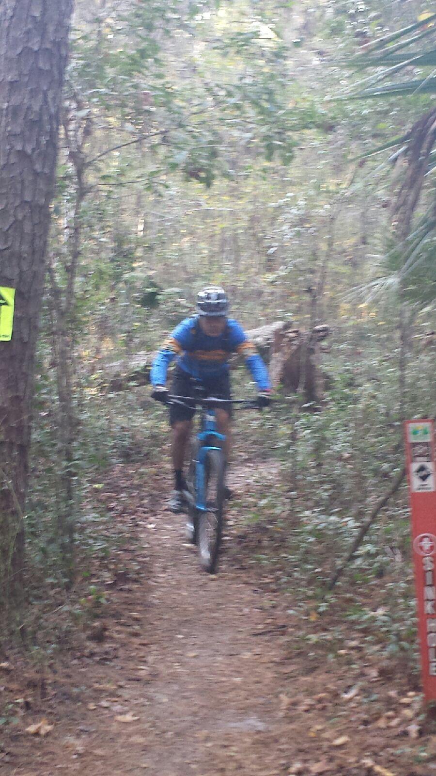 A person in a blue and yellow cycling jersey rides a mountain bike along a narrow trail in a dense, wooded area. Trees and underbrush surround the trail, while a caution sign is visible in the foreground. Santos mountain bike trail.