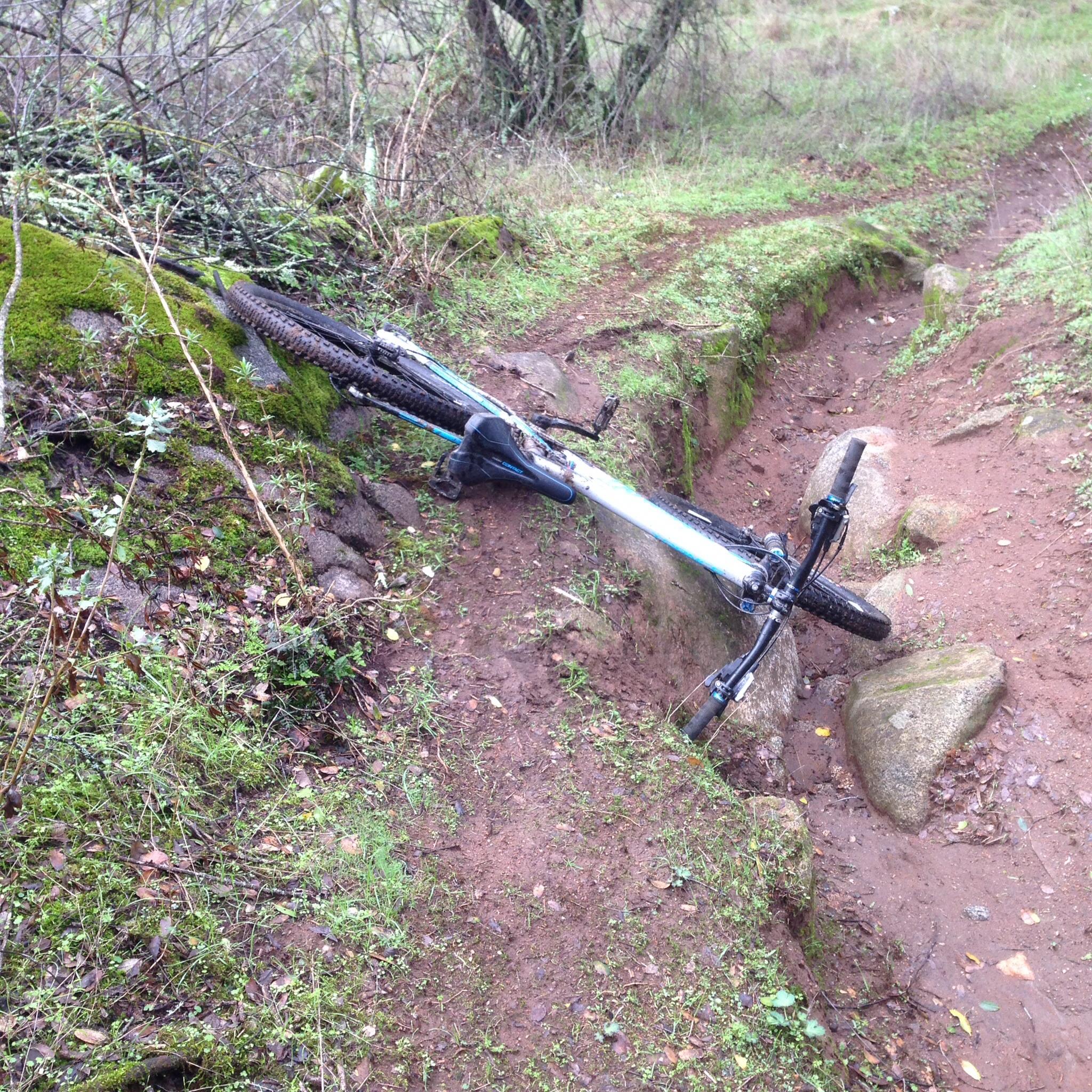A mountain bike lies on its side in a narrow, muddy trail surrounded by green grass and moss. The bike is partially in a shallow ditch, with rocks and vegetation visible nearby. Granite Bay Trail mountain bike trail.