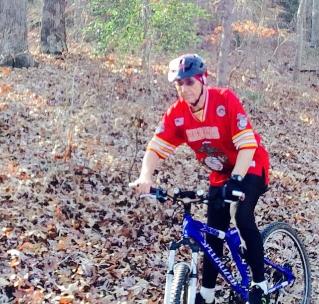 An older individual riding a blue mountain bike through a wooded area covered in autumn leaves, wearing a red sports jersey and a protective helmet. Cedarville State Forest mountain bike trail.