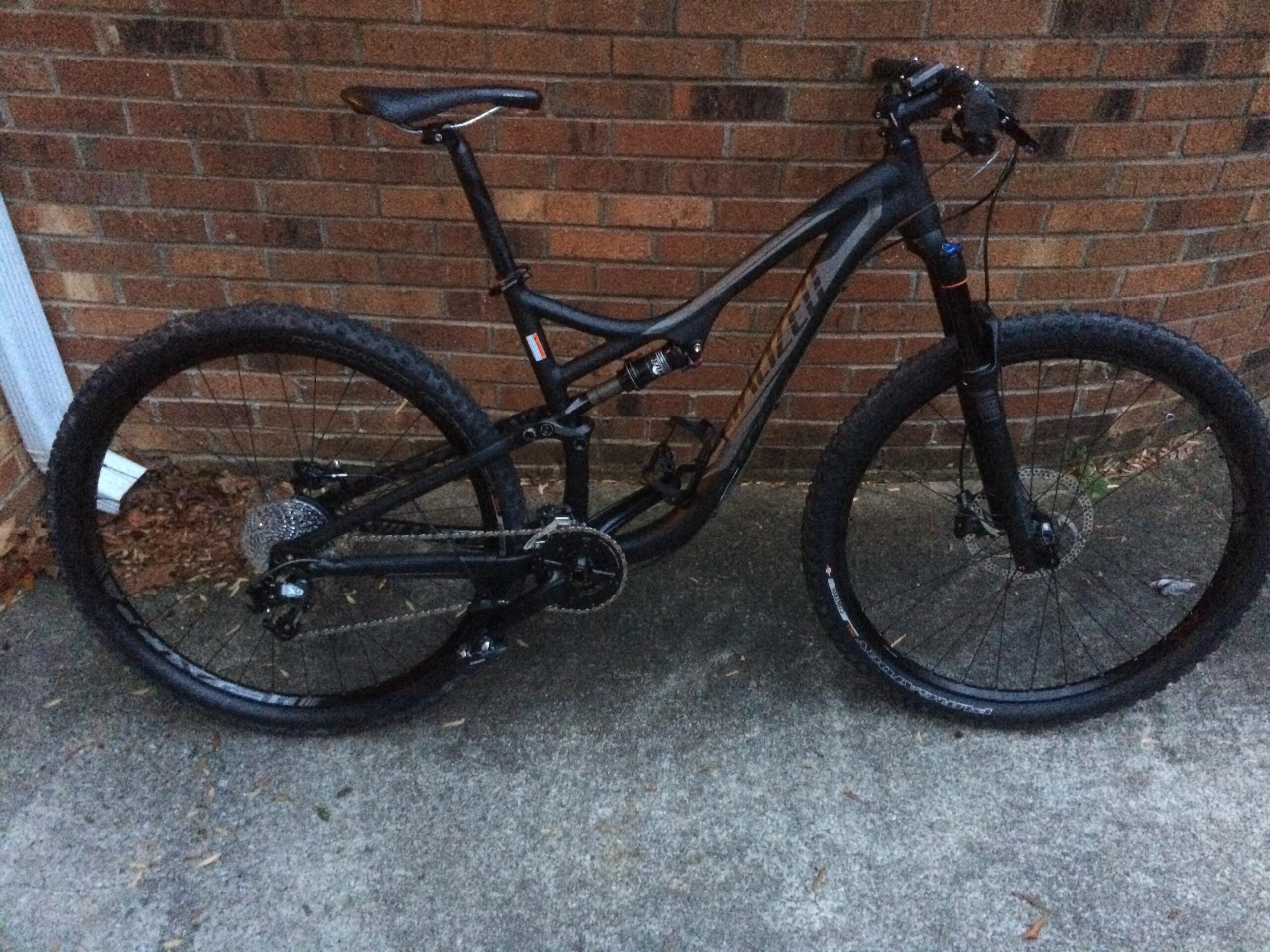 Specialized Stumpjumper FSR Comp 29er: A black mountain bike positioned against a brick wall, featuring thick tires and a modern frame design. The bike is equipped with a front suspension fork and a rear derailleur. The ground is paved, and there are some fallen leaves nearby.