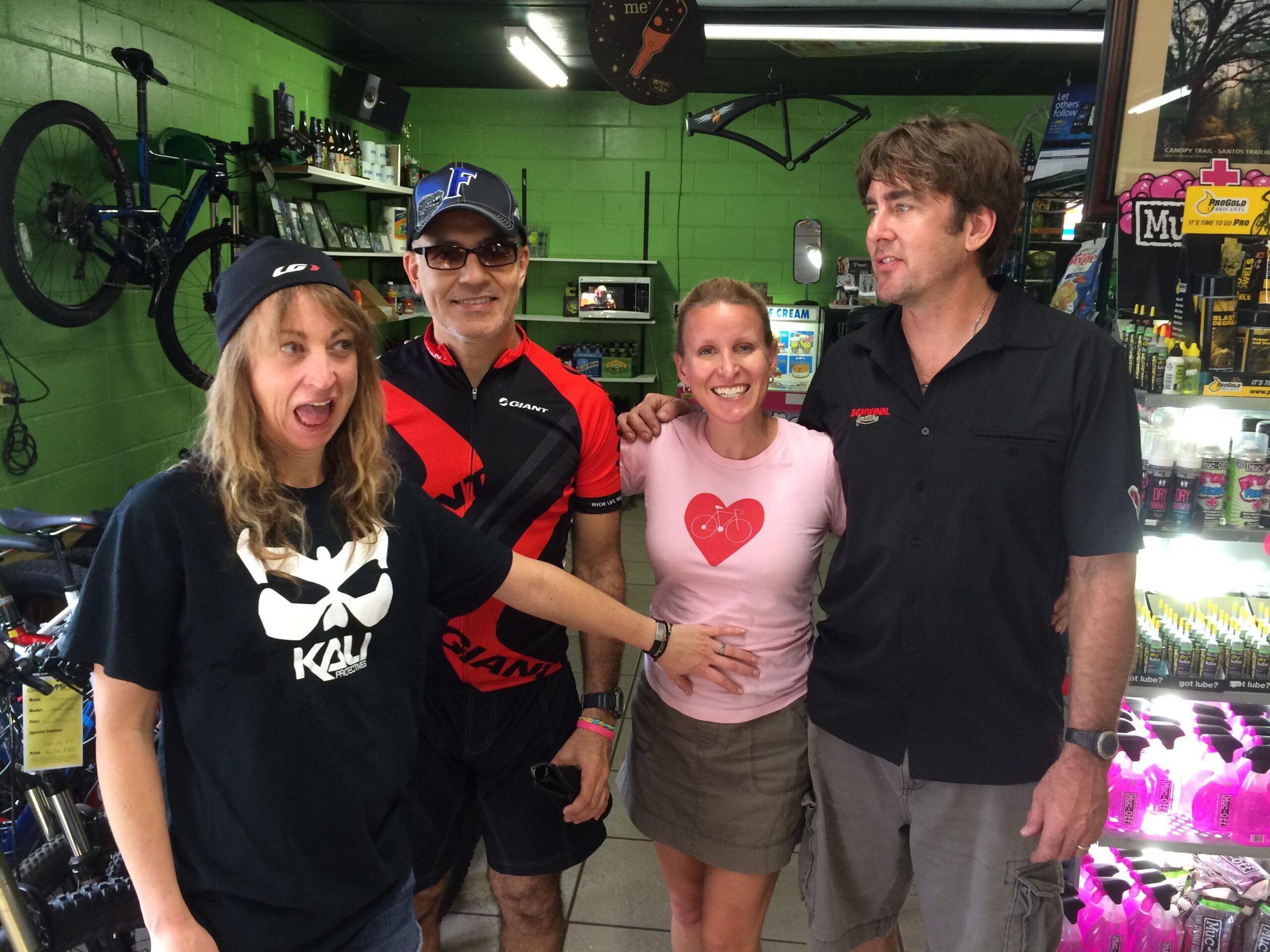 A group of four people poses together in a bicycle shop. The woman on the left makes a playful face, wearing a black shirt with a graphic design and a beanie. Next to her, a man in a red and black cycling jersey stands with a friendly smile. The woman in the center wears a pink shirt with a bicycle heart graphic and looks happy, while the man on the right, dressed in a black shirt, stands proudly beside her. The background features bicycles and various biking accessories displayed on green walls. Pine Tree Loop mountain bike trail.