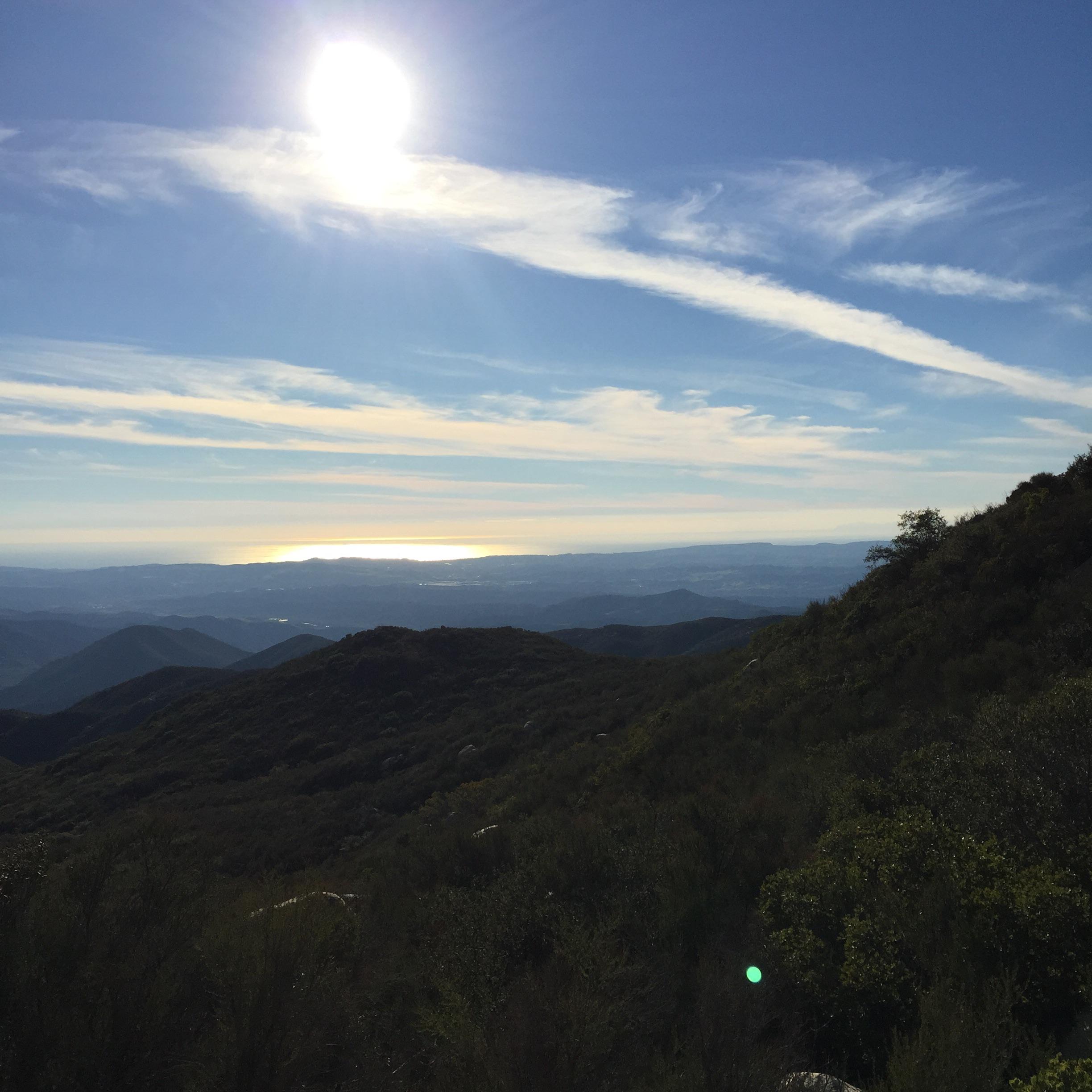 A panoramic view of rolling green hills under a bright sun, with a backdrop of a clear blue sky and wispy clouds. In the distance, the shimmering coastline can be seen, highlighting the contrast between the mountains and the ocean. San Juan Trail mountain bike trail.