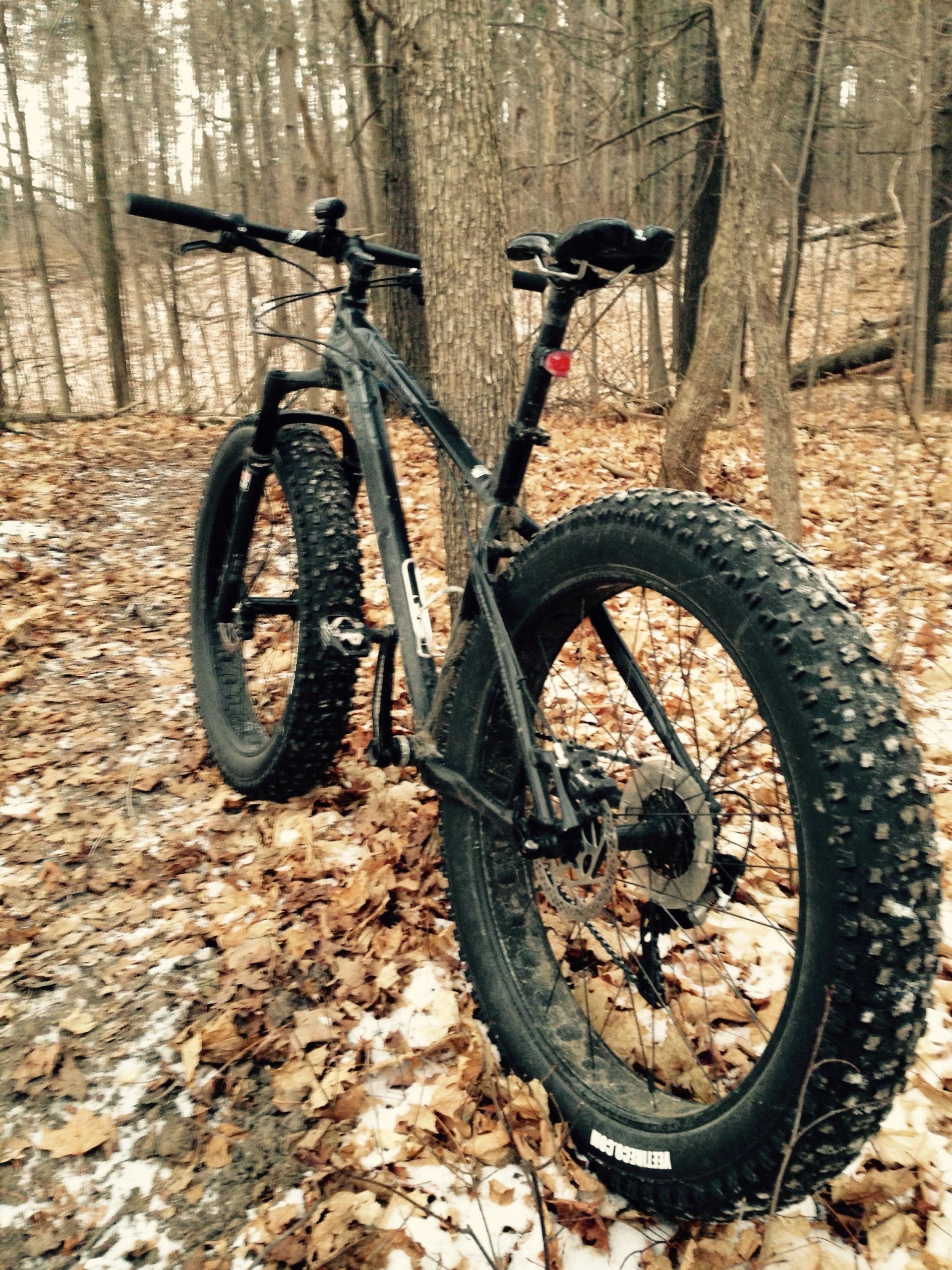 A black fat tire bike parked on a muddy trail surrounded by trees and fallen leaves, with patches of snow visible on the ground. The bike features wide, knobby tires designed for off-road terrain. Coulson's Hill mountain bike trail.