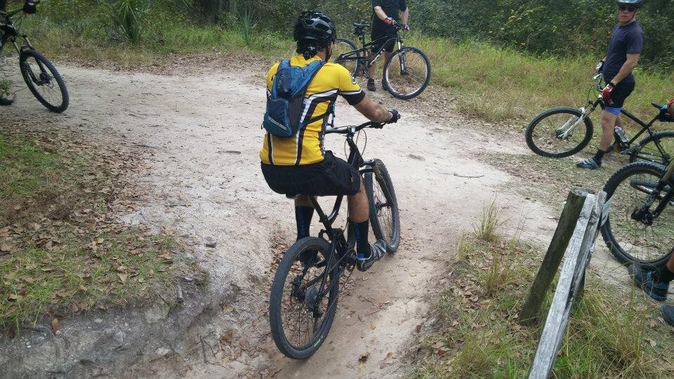 A group of mountain bikers gathered on a dirt trail in a forested area. One cyclist is positioned on a black bike, wearing a yellow cycling jersey and a backpack, while others are seen in the background on their bikes. The terrain is surrounded by greenery and natural elements, suggesting an outdoor recreational setting. Alafia River State Park mountain bike trail.