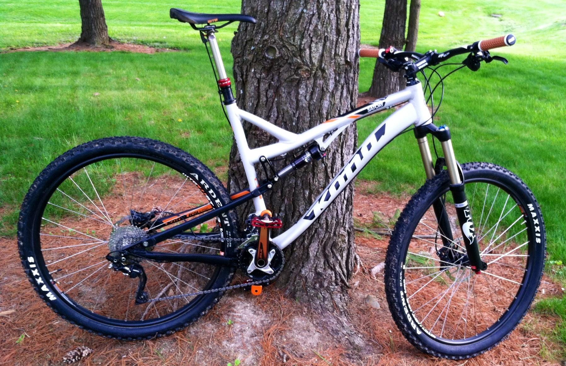 Kona Satori: A mountain bike with a white and black frame is leaning against a tree in a green grassy area. The bike features large knobby tires, a dropper seat post, and various components that indicate it's designed for off-road riding. Pine needles and small stones are visible on the ground around the bike.