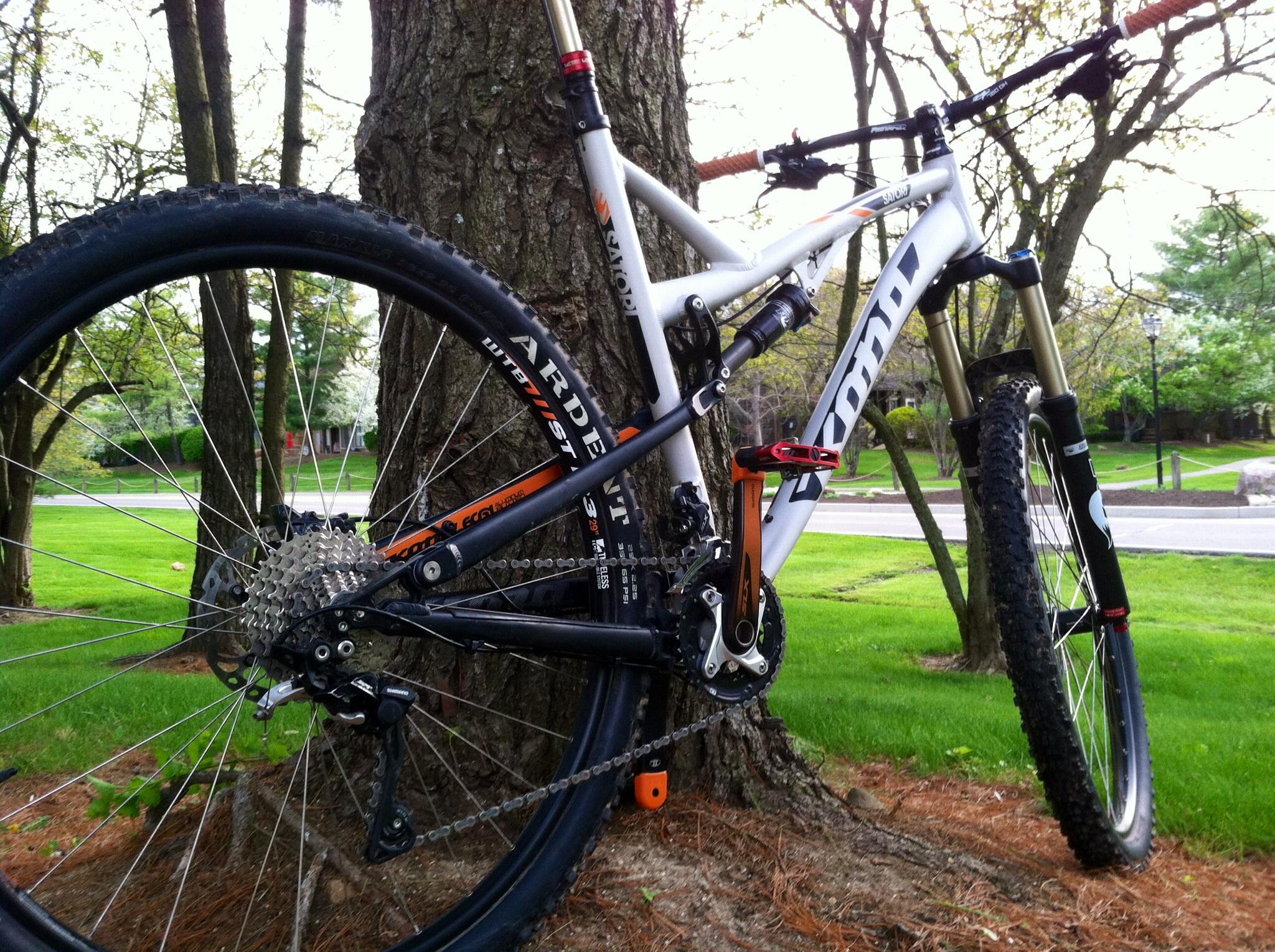 Kona Satori: A mountain bike leaning against a tree, featuring thick tires and a detailed drivetrain. The bike has a white and black color scheme with orange accents, surrounded by green grass and trees in a park setting.