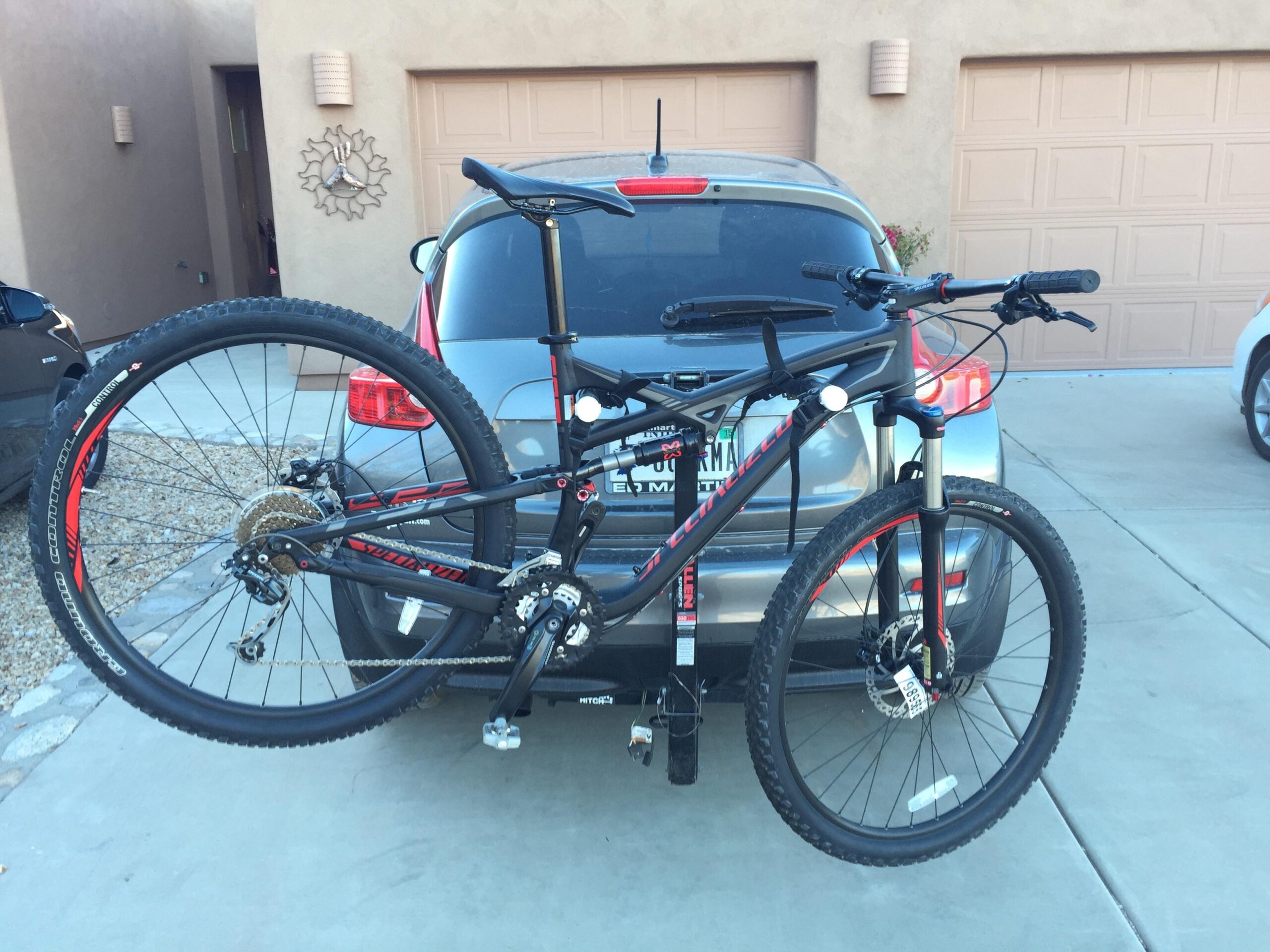 Specialized Camber 29: A mountain bike mounted on the back of a car, with the bike's front wheel elevated and the rear wheel closer to the vehicle. The background shows a light-colored house and a garage door, along with some outdoor decor. The scene is set in a driveway.