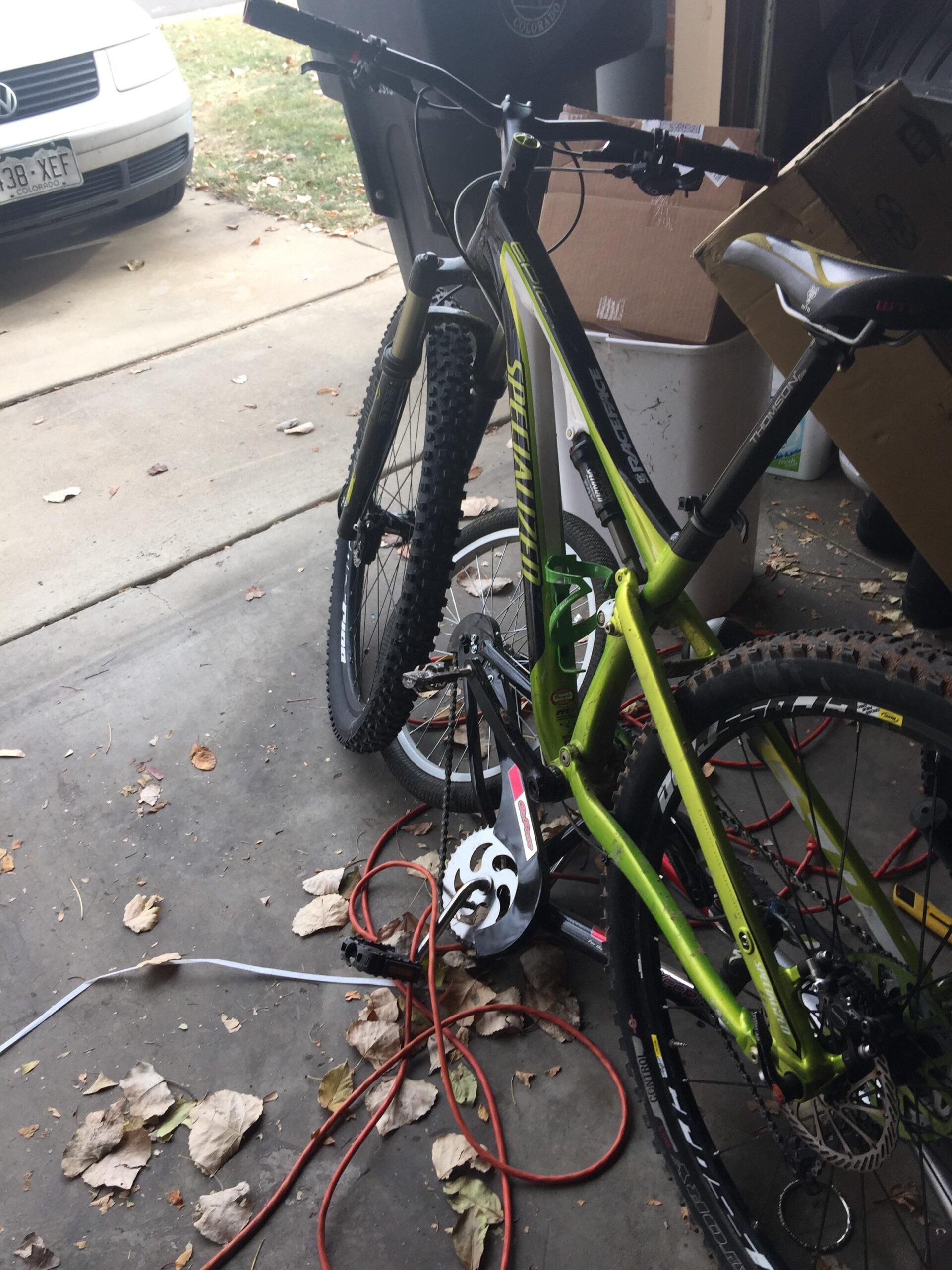 Specialized Epic Comp 29er: A green mountain bike is leaning against a garage wall with visible tools and cables on the floor. Brown leaves are scattered around the concrete surface. In the background, a white car is parked on the driveway, partially visible. Cardboard boxes are stacked nearby.
