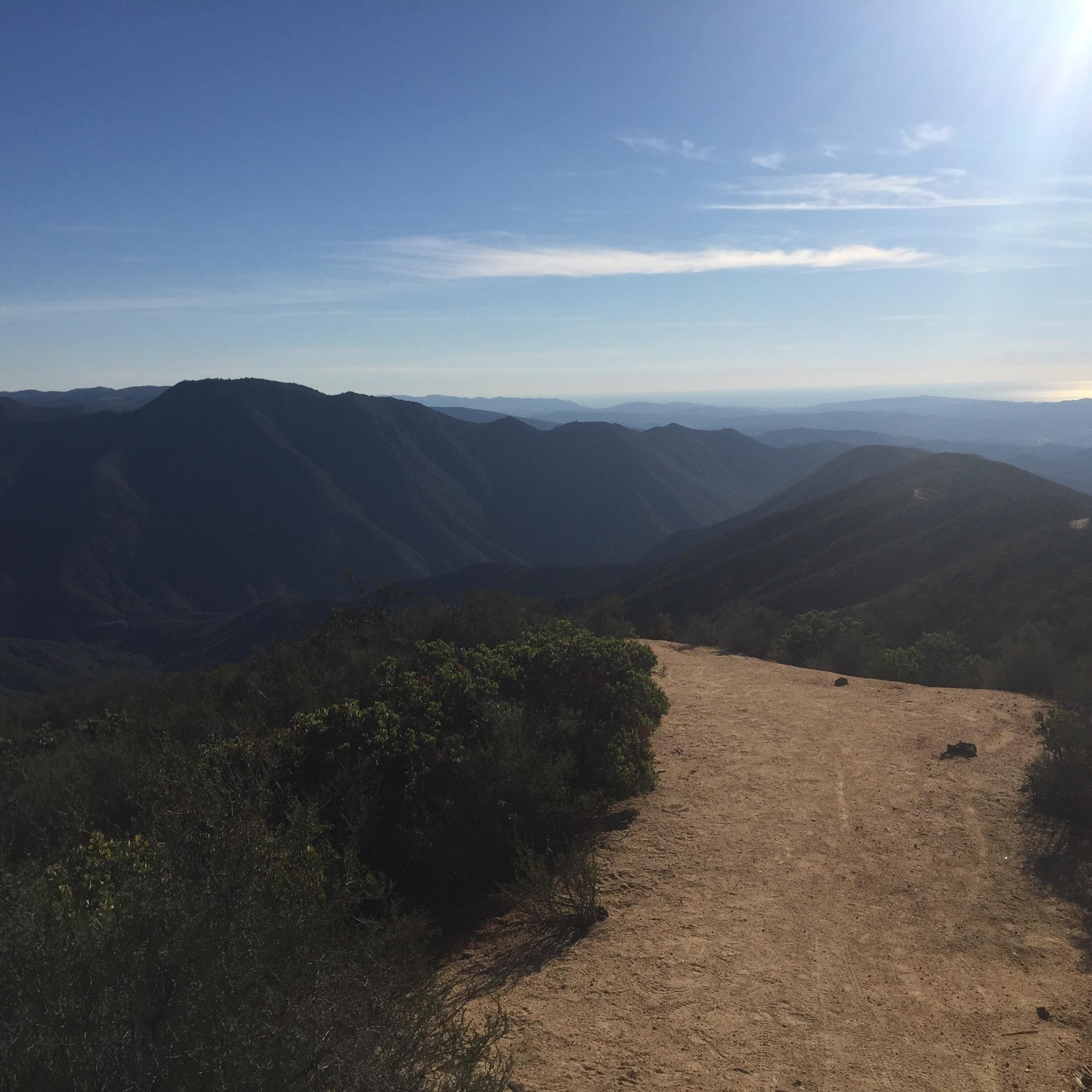 A scenic view of a mountain landscape under a clear blue sky, featuring rolling hills and distant mountains. A dirt path winds through the foreground, bordered by green shrubs. The sunlight casts a warm glow on the terrain, creating a peaceful and inviting atmosphere. San Juan Trail mountain bike trail.