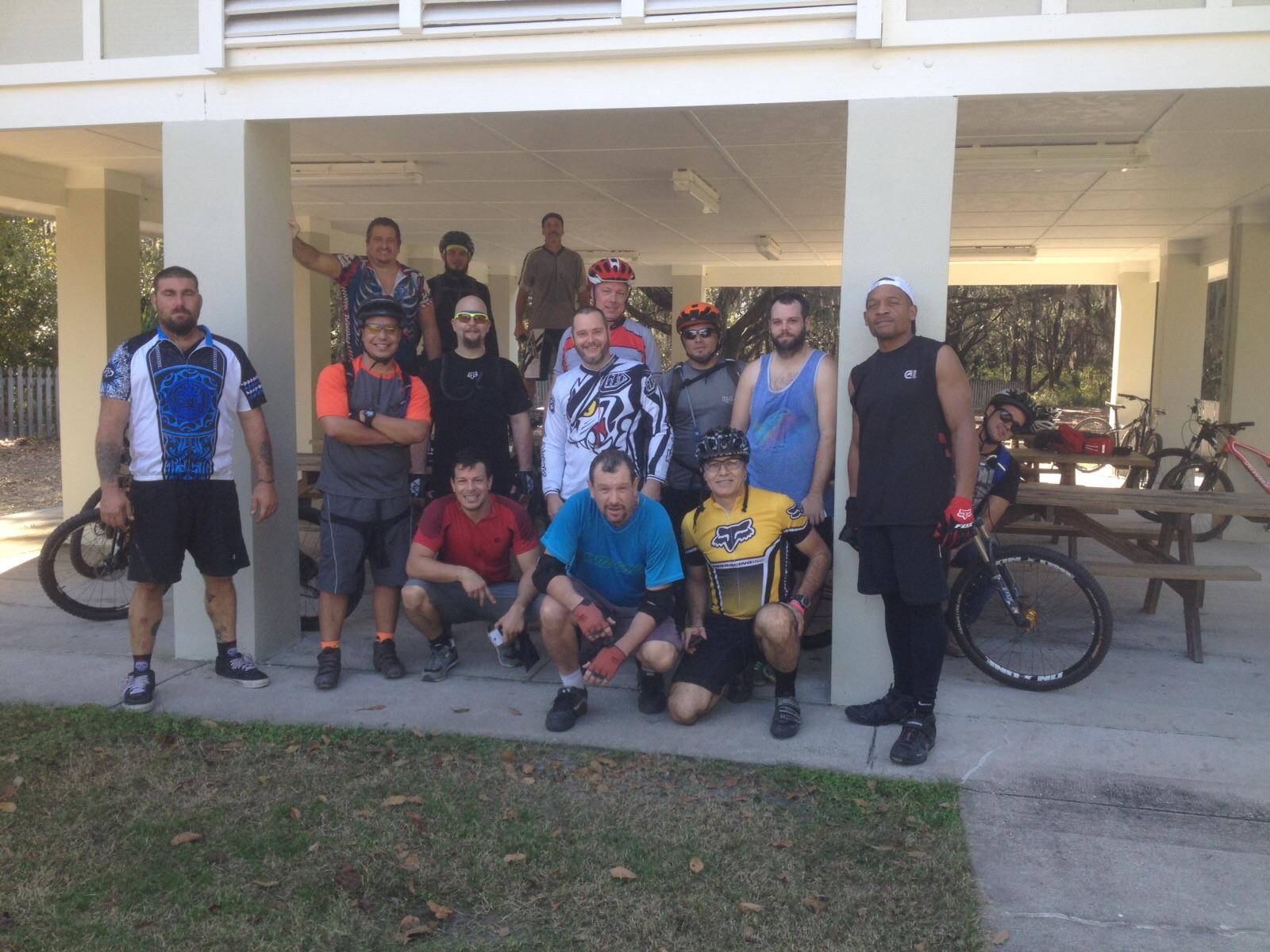 A group of men in cycling attire pose for a photo outdoors, with mountain bikes partially visible in the background. They are gathered under a shaded area, with some standing and others crouching. The setting appears to be a park or recreation area, with trees in the background and picnic tables nearby. Alafia River State Park mountain bike trail.